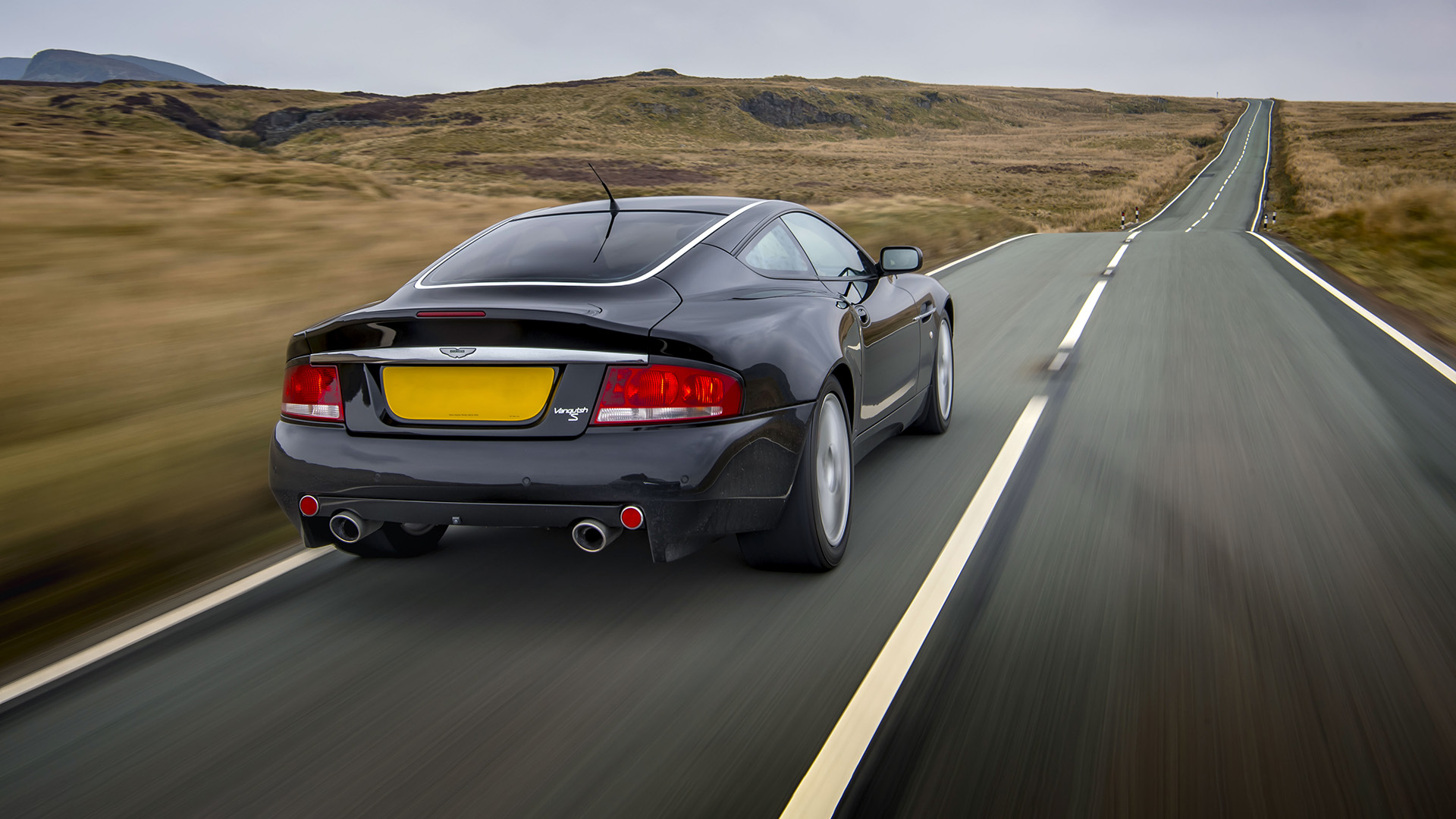 Rear view of a sleek black Aston Martin Vanquish S cruising on a scenic country road. 