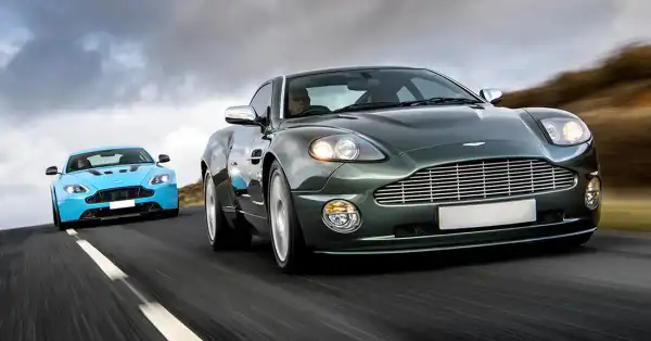 Two Aston Martins driving on a road with a scenic countryside background.