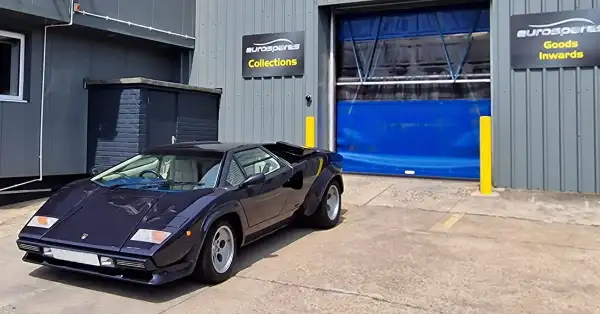 Blue Lamborghini Countach parked outside the Eurospares warehouse with customer collection and goods inward signs on the wall.